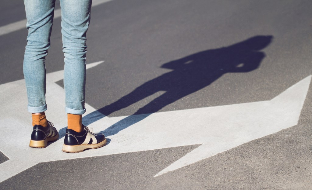 Close up side view of person wearing black shoes and blue jeans standing on a street with arrow signs pointing in different directions