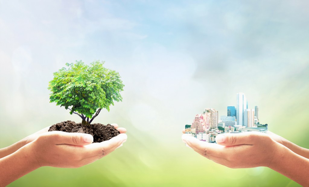 Two human hand holding heart shape of tree and big city over blurred green nature background