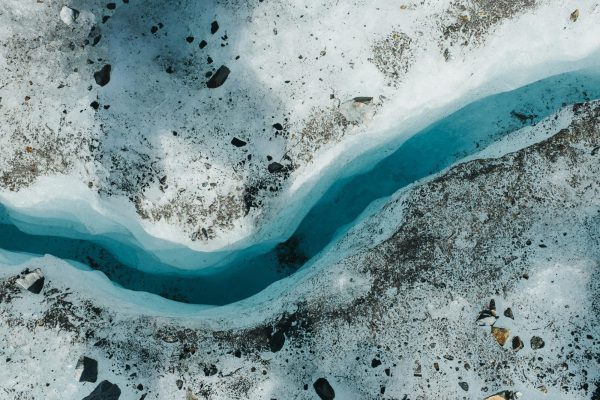 Overhead view of a glacier surface, with a narrow blue meltwater stream winding through white ice scattered with rocks and sediment.